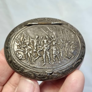 May include: Oval silver-toned metal box with a hinged lid. The lid features a detailed relief scene of figures in historical attire, possibly soldiers, with flags and weapons. The box has an ornate, floral-like border.