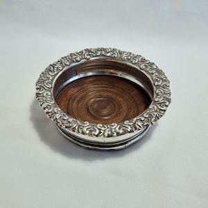 May include: A silver-toned coaster with an ornate, scrolling design around the rim. The coaster's interior features a dark brown, circular wooden base. The coaster is designed to protect surfaces from drink condensation.