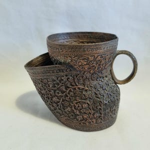 Puede incluir: Una taza de bronce de estilo antiguo con asa y una taza más pequeña en su interior. La superficie está intrincadamente tallada con patrones florales, mostrando una estética vintage. La taza se encuentra sobre un fondo blanco liso.