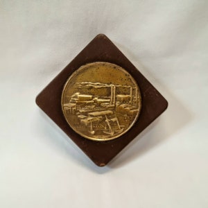 Può includere: Una scatola quadrata in legno marrone con una placca circolare in metallo dorato che presenta un'immagine in rilievo di un treno con fumo che esce dal camino. Il treno è circondato da edifici e alberi.