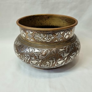 May include: A round, brass bowl with intricate silver inlay. The bowl features a decorative pattern of floral motifs and Arabic calligraphy.