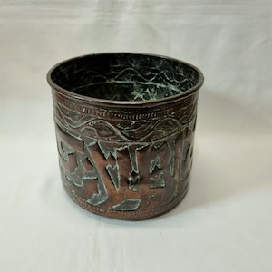 Könnte beinhalten: Ein Kupfertopf mit einem komplizierten Prägemuster. Der Topf hat eine runde Form und einen leicht ausgestellten Rand. Das Muster zeigt ein sich wiederholendes Muster aus stilisierten Blättern und Blumen.