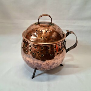 Peut inclure: Une marmite en cuivre avec un couvercle et une poignée. La marmite a un corps arrondi avec de petits trous et repose sur trois petits pieds. Le couvercle a également de petits trous et une petite poignée. La marmite a une texture martelée.