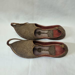 Pu&ograve; includere: Un paio di pantofole antiche in pelle marrone ornate, con punta e un anello sul tallone. Le pantofole presentano un motivo floreale in rilievo e un interno rosso. Le suole sono marrone scuro.