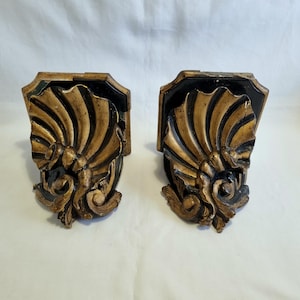 May include: Pair of ornate, vintage wall sconces. Each sconce features a black rectangular base with a gold, fan-shaped design and decorative scrollwork. The sconces are made of a dark material and have a distressed finish.