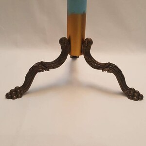 May include: A gold-painted metal stand with three ornate legs. The legs are curved and have a decorative design. The stand is on a white surface.