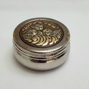 Puede incluir: Una caja redonda de plata para alhajas con una tapa que presenta un diseño en relieve de tres ángeles con alas, rodeados de nubes, en oro.