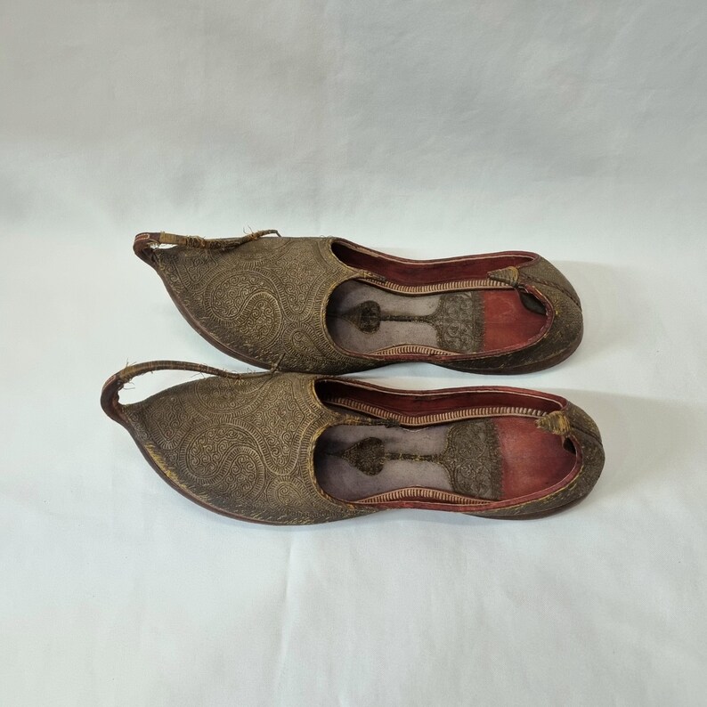 Pu&ograve; includere: Un paio di pantofole antiche in pelle marrone ornate, con punta e un anello sul tallone. Le pantofole presentano un motivo floreale in rilievo e un interno rosso. Le suole sono marrone scuro.