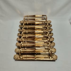 Puede incluir: Una pila de tiradores dorados para armarios. Cada tirador tiene un asa curvada y placas decorativas en los extremos. Los tiradores están dispuestos en una pila ordenada y organizada, lo que sugiere un juego para un proyecto de mobiliario o una renovación. El herraje tiene una superficie brillante y reflectante.