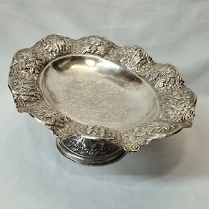 May include: A silver serving dish with an ornate design featuring grapevines and leaves. The dish has a raised base and a scalloped edge.