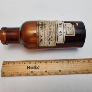 以下が含まれることがあります： 「Pulv. Jalapa」と「The British Drug House」と書かれたラベルが付いた、アンティークの茶色のガラス瓶。細い首と「B.P. 1932」と書かれたラベルが付いた円筒形。木製の定規が瓶の隣にあります。