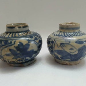 May include: Two small, antique ceramic vases with a blue and cream floral design. The vases have a rounded body and a short neck. The blue floral pattern is hand-painted on a cream background. These decorative vases are likely for display.