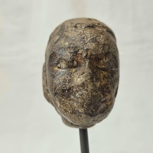 Puede incluir: Busto desgastado de una cabeza humana, de estilo antiguo. La escultura es de color marrón apagado con textura y detalles visibles, incluyendo ojos cerrados y una nariz y boca definidas. La cabeza está montada sobre una varilla de metal oscuro.