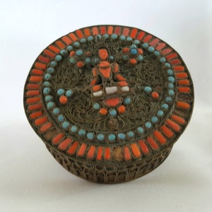 May include: A round, silver-coloured box with a decorative lid. The lid is adorned with intricate filigree work and is inlaid with turquoise and coral stones. The box is approximately 7.5 cm in diameter.