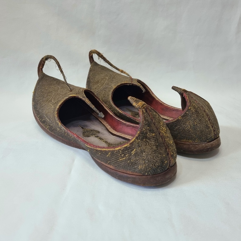Pu&ograve; includere: Un paio di scarpe slip-on marroni ornate, con una punta curva e un anello sul tallone. Le scarpe presentano un motivo in rilievo intricato e un interno rosso. Le suole sono marrone scuro e le scarpe sembrano essere realizzate in tessuto strutturato.