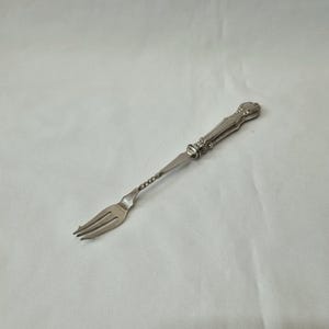 May include: A silver fork with a decorative handle and three tines. The handle has a twisted design and ornate detailing. The fork is likely vintage or antique, and is set against a white background.