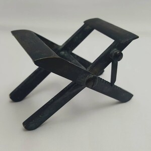 May include: A small, dark metal folding chair with an X-shaped base and curved seat. The chair has a simple design with a hinge and screw mechanism. The overall color is a dark, aged metal, suggesting it is vintage or antique.