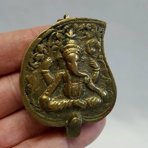 May include: A brass pendant depicting a detailed relief of the Hindu deity Ganesha, seated in a meditative pose with his trunk raised. The pendant is leaf-shaped and features intricate floral designs around the edges.