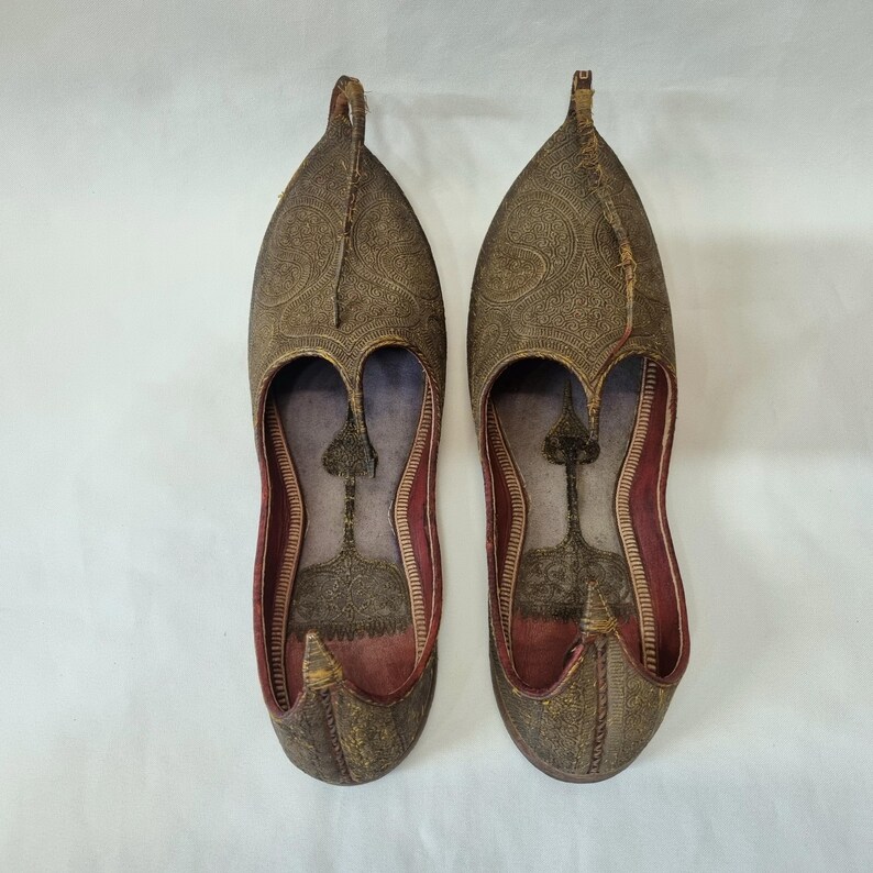 Pu&ograve; includere: Un paio di scarpe ornate in pelle marrone con intricati motivi in rilievo. Le scarpe hanno una punta affusolata e una tomaia curva, con interni rossi. Le scarpe sono probabilmente fatte a mano e hanno un design tradizionale.