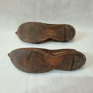 Pu&ograve; includere: Due antiche scarpe in pelle marrone, suole rivolte verso l'alto, che mostrano usura e dettagli delle cuciture. Le scarpe hanno una punta arrotondata e un piccolo tacco. La pelle appare invecchiata e strutturata, suggerendo che siano vintage o fatte a mano.