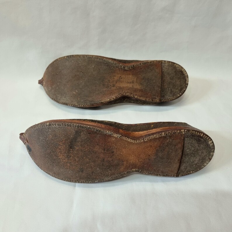 Pu&ograve; includere: Due antiche scarpe in pelle marrone, suole rivolte verso l'alto, che mostrano usura e dettagli delle cuciture. Le scarpe hanno una punta arrotondata e un piccolo tacco. La pelle appare invecchiata e strutturata, suggerendo che siano vintage o fatte a mano.