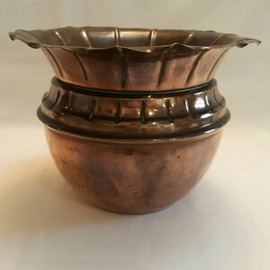 May include: A copper-toned planter with a decorative, scalloped rim and a rounded base. The planter features a two-part design, with a patterned band around the middle. The metal has a warm, aged patina, suggesting it is vintage or antique.