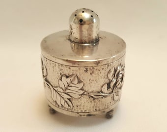 Op de afbeelding: Een kleine, antieke zilveren zoutstrooier met een getextureerd oppervlak en een bloemenreliëf. De strooier heeft een cilindrisch lichaam, een plat deksel en een kleine, geperforeerde bovenkant. Hij staat op drie kleine voetjes.