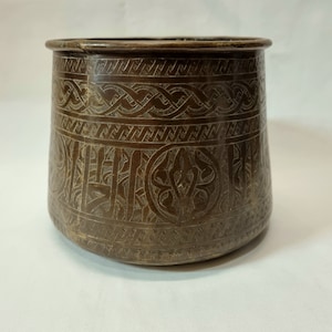 Könnte beinhalten: Eine braune Metallschale mit aufwendigen gravierten Mustern. Die Schale hat einen breiten, abgerundeten Boden und einen leicht ausgestellten Rand.