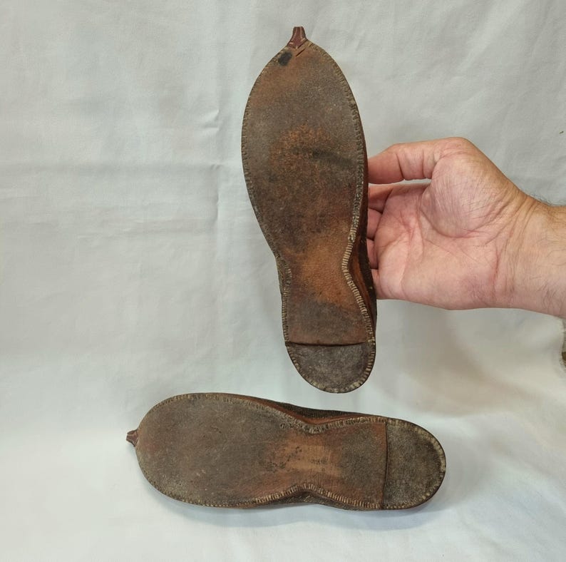 Pu&ograve; includere: Due suole di scarpe antiche in pelle marrone, che mostrano usura e cuciture. Le suole sono di forma ovale con una punta affusolata e un piccolo tacco. La pelle ha un aspetto vissuto, che suggerisce et&agrave; e utilizzo.