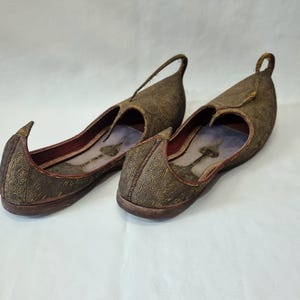 Pu&ograve; includere: Un paio di scarpe marroni ornate, senza lacci, con punta e anello sul tallone. Le scarpe hanno un design a fantasia e un interno rosso. Le suole sono marrone scuro.