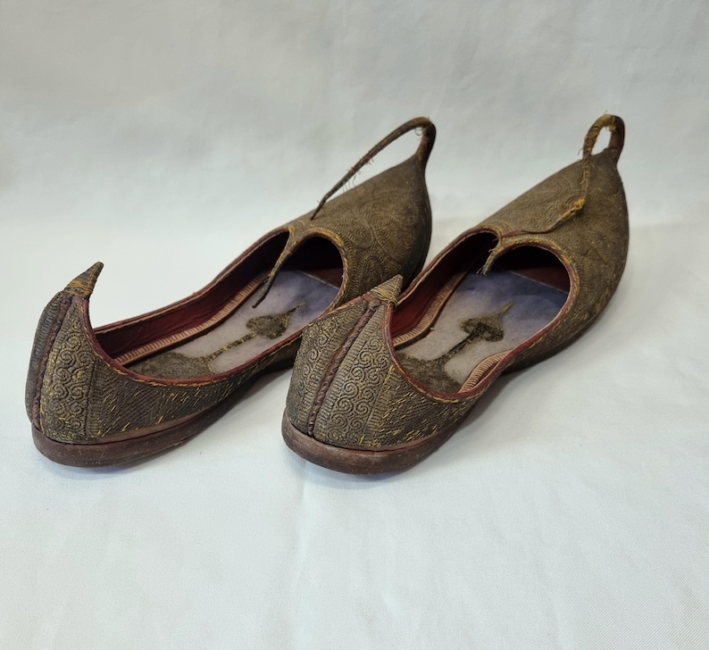 Pu&ograve; includere: Un paio di scarpe marroni ornate, senza lacci, con punta e anello sul tallone. Le scarpe hanno un design a fantasia e un interno rosso. Le suole sono marrone scuro.