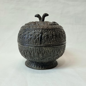 Op de afbeelding: Een kleine, ronde, zwarte metalen doos met een ingewikkeld, gegraveerd ontwerp. De doos heeft een deksel met een decoratieve knop.