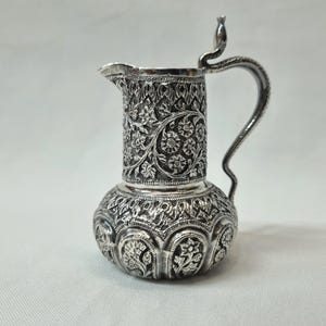 Può includere: Un'anfora d'argento con un designo floreale ornato. L'anfora ha un corpo rotondo e un beccuccio con un manico curvo. L'anfora è decorata con intricati motivi floreali.