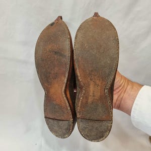 Pu&ograve; includere: Un paio di scarpe in pelle marrone usurate, suole rivolte verso l'alto, che mostrano segni di utilizzo. Le scarpe hanno un design a punta e sono tenute da una mano su sfondo bianco. Le suole mostrano cuciture.