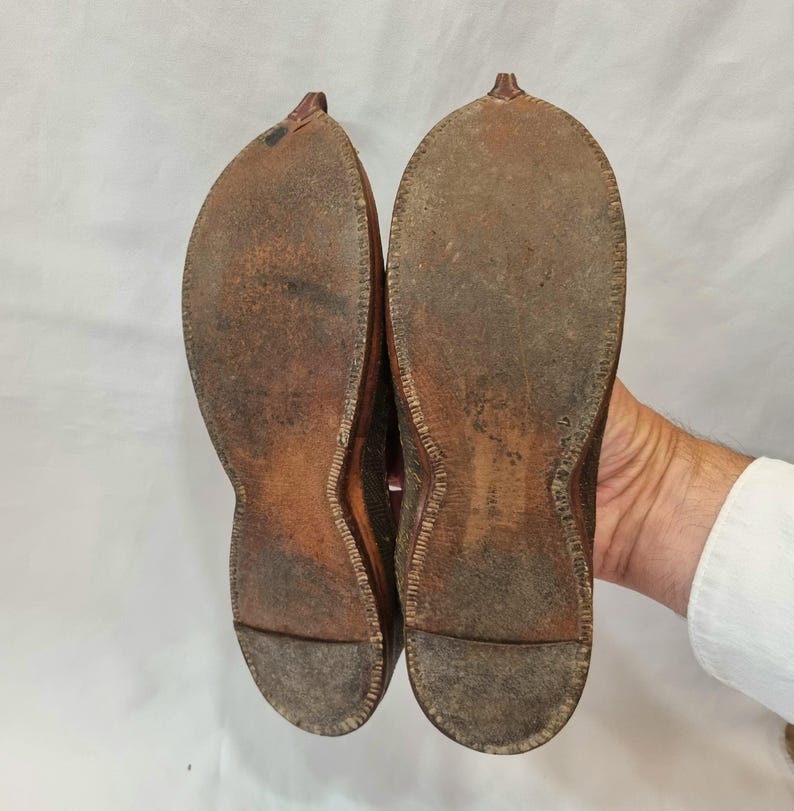 Pu&ograve; includere: Un paio di scarpe in pelle marrone usurate, suole rivolte verso l'alto, che mostrano segni di utilizzo. Le scarpe hanno un design a punta e sono tenute da una mano su sfondo bianco. Le suole mostrano cuciture.
