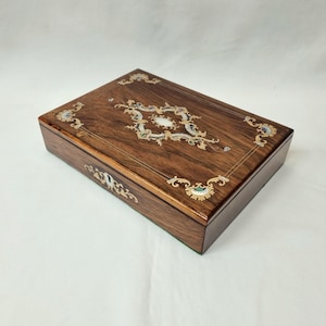 May include: A wooden box with a hinged lid, inlaid with mother-of-pearl and gold accents. The box has a decorative floral design on the top.