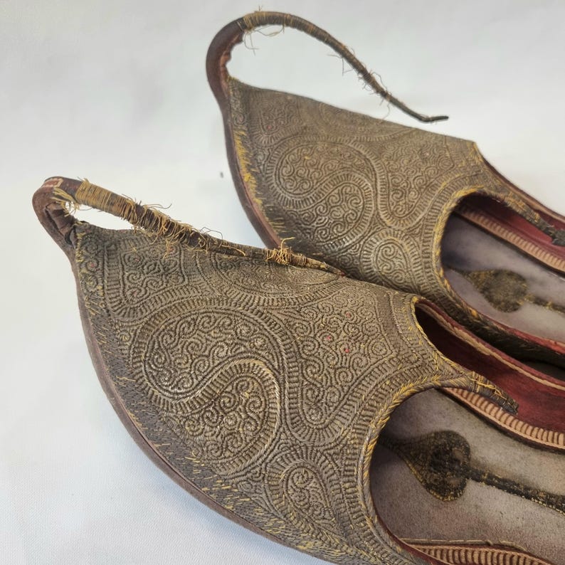 Pu&ograve; includere: Un paio di pantofole antiche in pelle marrone con intricati motivi paisley in rilievo. Le pantofole hanno una punta a punta e un cinturino curvo e ad anello sul tallone. L'interno &egrave; foderato in tessuto rosso.
