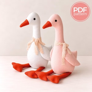 May include: Two plush goose toys, one white and one light pink, each featuring a bright orange beak and orange webbed feet. Both geese have small black eyes and are adorned with a delicate, light-coloured lace ribbon tied around their necks.