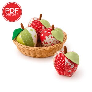 May include: A PDF pattern for fabric apples featuring three stuffed apples in a woven wicker basket and one separate fabric apple. The apples are crafted from various red, white, and green patterned fabrics, each topped with a brown stem and a light green leaf.