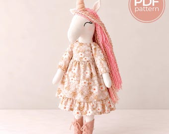 May include: A fabric unicorn doll with a spiral horn, long pink yarn hair, and embroidered eyes. The doll wears a beige dress with a white floral pattern and a ruffled hem, paired with light brown lace-up boots. Includes a PDF pattern label.