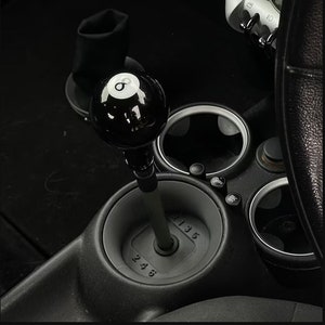 Könnte beinhalten: Schwarzer 8-Ball-Schalthebelknauf für ein Auto. Der Knauf besteht aus glänzendem schwarzem Material und hat eine weiße 8 darauf. Der Knauf ist an einem silbernen Schalthebel befestigt. Der Schalthebel ist von einem schwarzen Schalthebelbalg umgeben, auf dem die Zahlen 1, 2, 3, 4, 5 und 6 aufgedruckt sind.