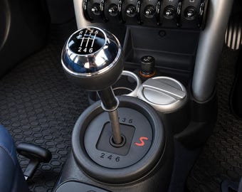 May include: A manual transmission gear shifter with a round, metallic knob featuring a black gear pattern insert. The base plate displays the numbers 1, 3, 5, 2, 4, 6 and a red letter S. The center console includes toggle switches and cup holders.