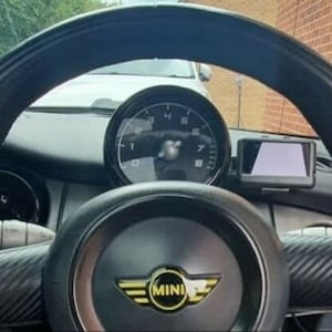 Op de afbeelding: Close-up van een zwart lederen stuurwiel met een zilveren Mini Cooper-logo en een zwart dashboard met een snelheidsmeter en een scherm.