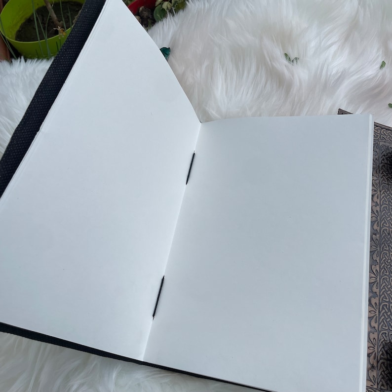 May include: An open blank notebook with black covers and black stitching. The notebook is lying open on a white fluffy surface.