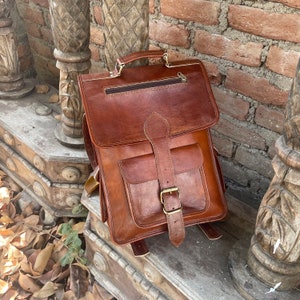 May include: A brown leather backpack with a flap closure, two front pockets, and adjustable straps. The backpack has a brass buckle and hardware.