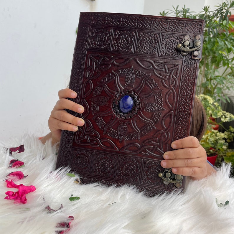 May include: A brown leather journal with intricate Celtic knotwork designs and a blue gemstone in the centre. The journal has a metal clasp and is closed.