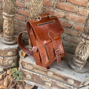 May include: A brown leather backpack with a flap closure and a buckle. The backpack has a large pocket on the front and a smaller pocket on the back. The backpack is sitting on a wooden surface.