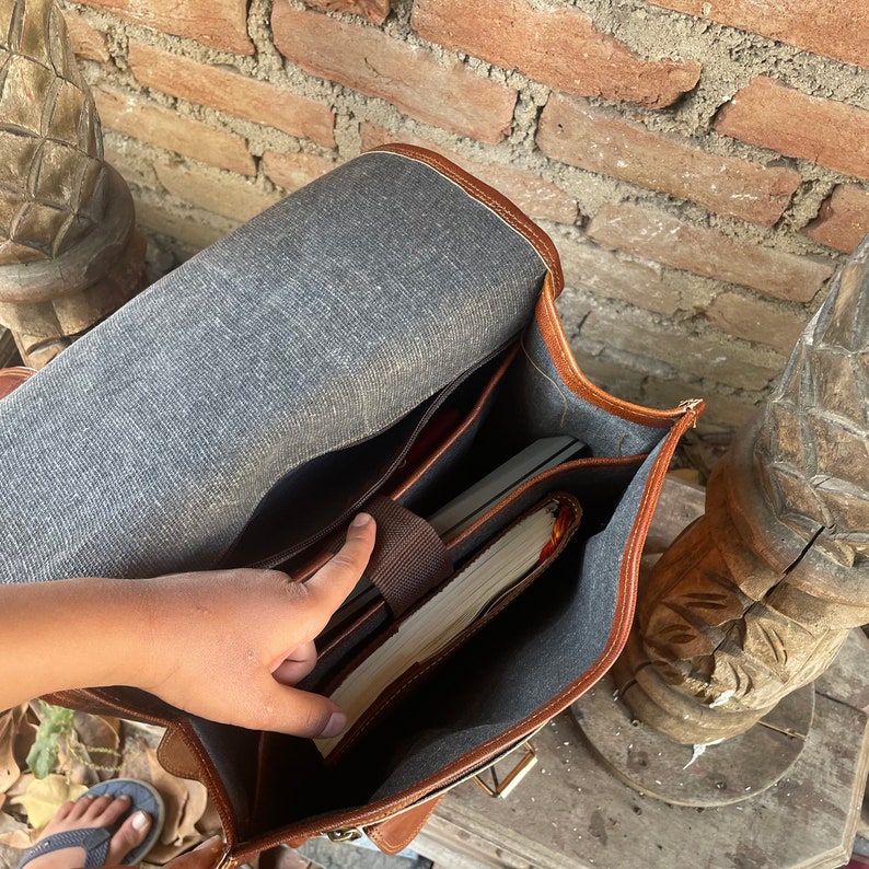 May include: A brown leather satchel bag with a gray fabric lining. The bag is open and has a few items inside, including a book and a notebook.