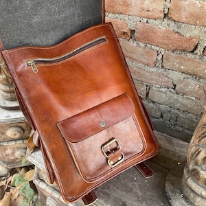 May include: A brown leather backpack with a large front pocket and a buckle closure. The backpack has a zippered top closure and a brown canvas lining.