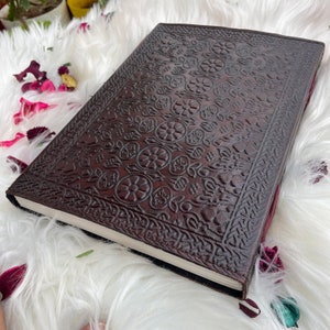 May include: A brown leather journal with an intricate embossed floral pattern. The journal has a blank page visible and is closed.